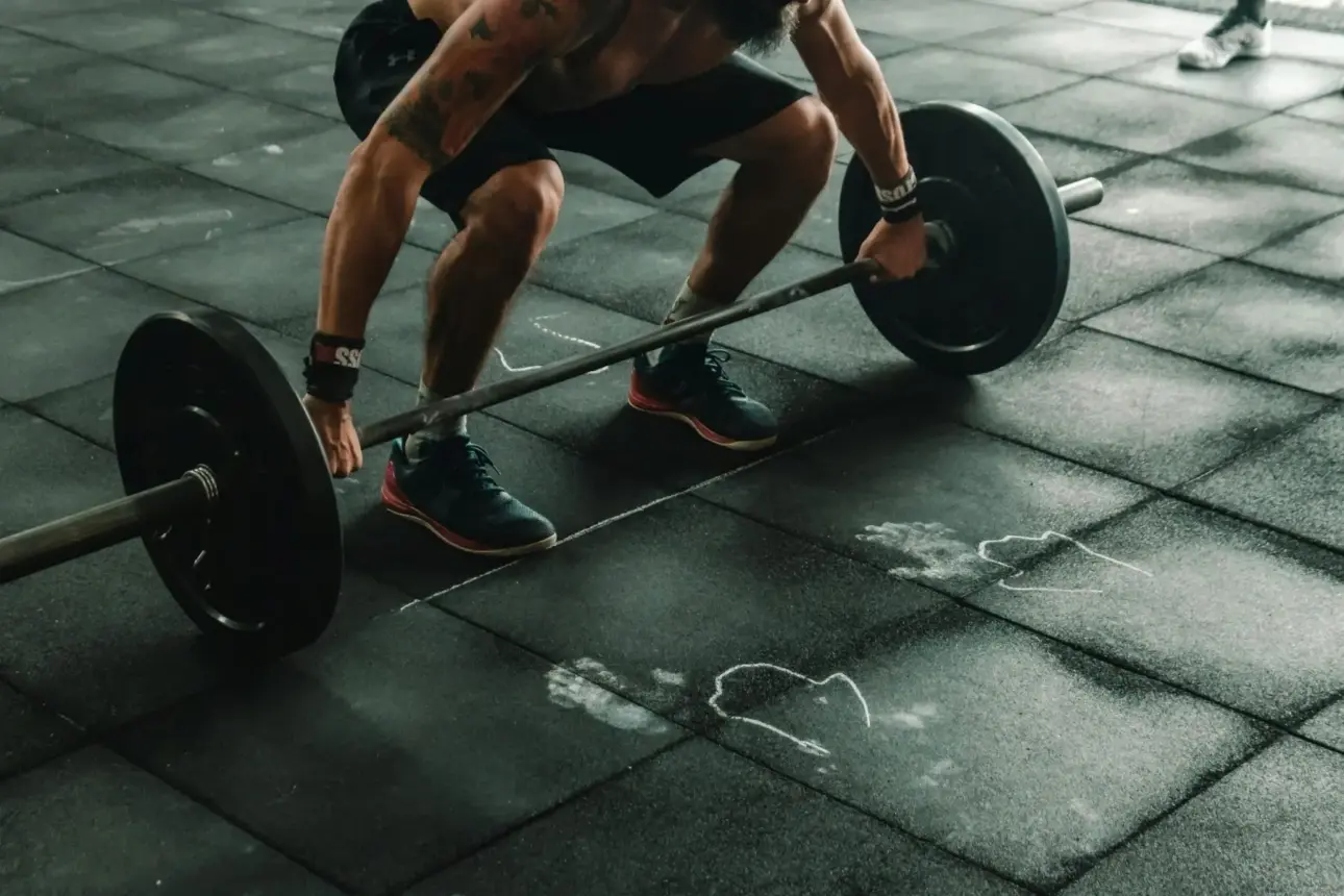 Homme saisissant une barre d’haltérophilie sur un sol de gym — préparation au soulevé de terre (deadlift), barre olympique/powerlifting acier haute résistance, équipement musculation pour force et puissance, séance de traction du bas du dos et jambes.