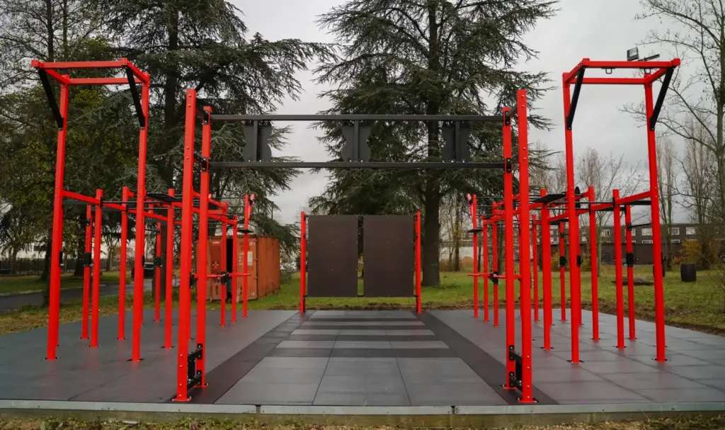 Dalles caoutchouc noires et grises posées en extérieur sous une structure de street workout rouge