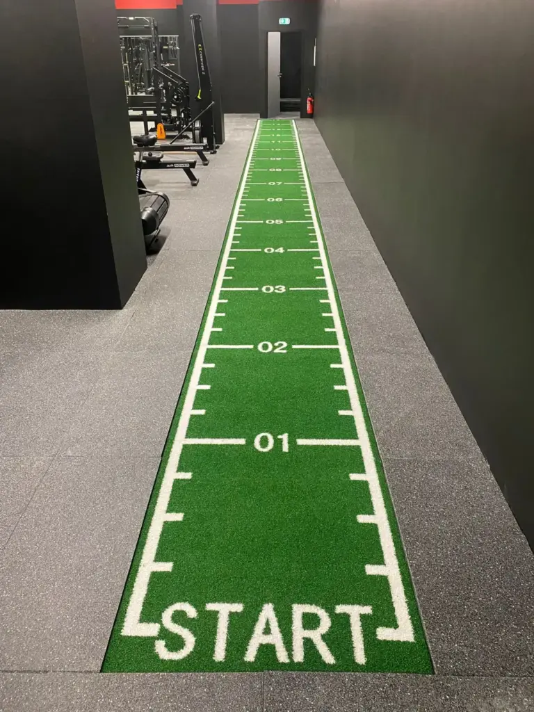 Piste en gazon synthétique vert graduée avec marquage START, posée le long d’un couloir de salle de sport
