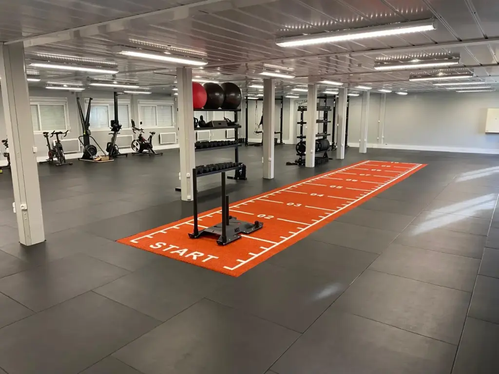 Brick-red synthetic turf sprint track with sled, laid in a gym on black rubber tiles